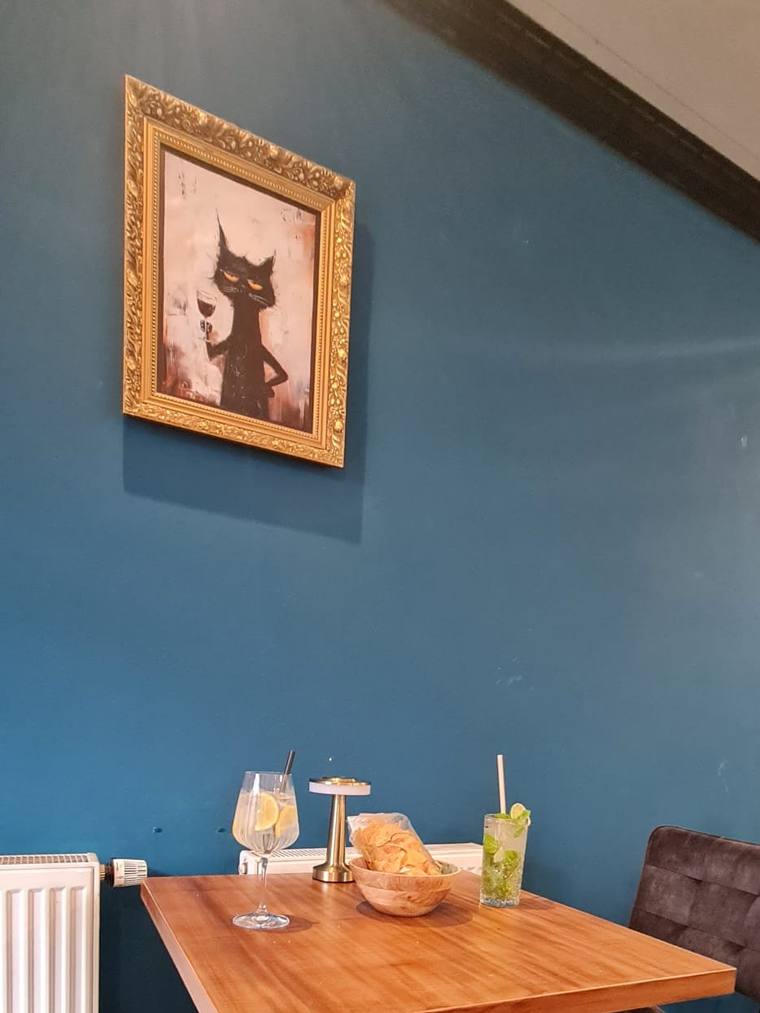 Table by a teal wall under the gold-framed cat painting, drinks on the table, chairs.