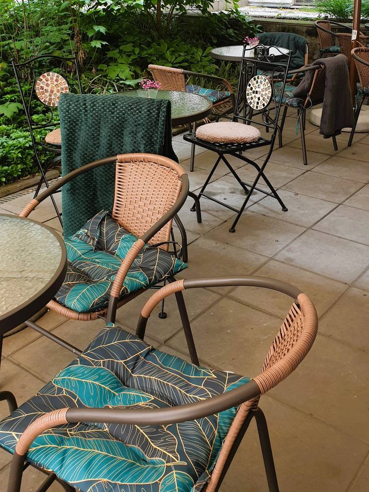 Courtyard terrace close-up, portrait: rattan chairs with teal leaf-print cushions, blankets, round tables, lush greenery and parasol above.